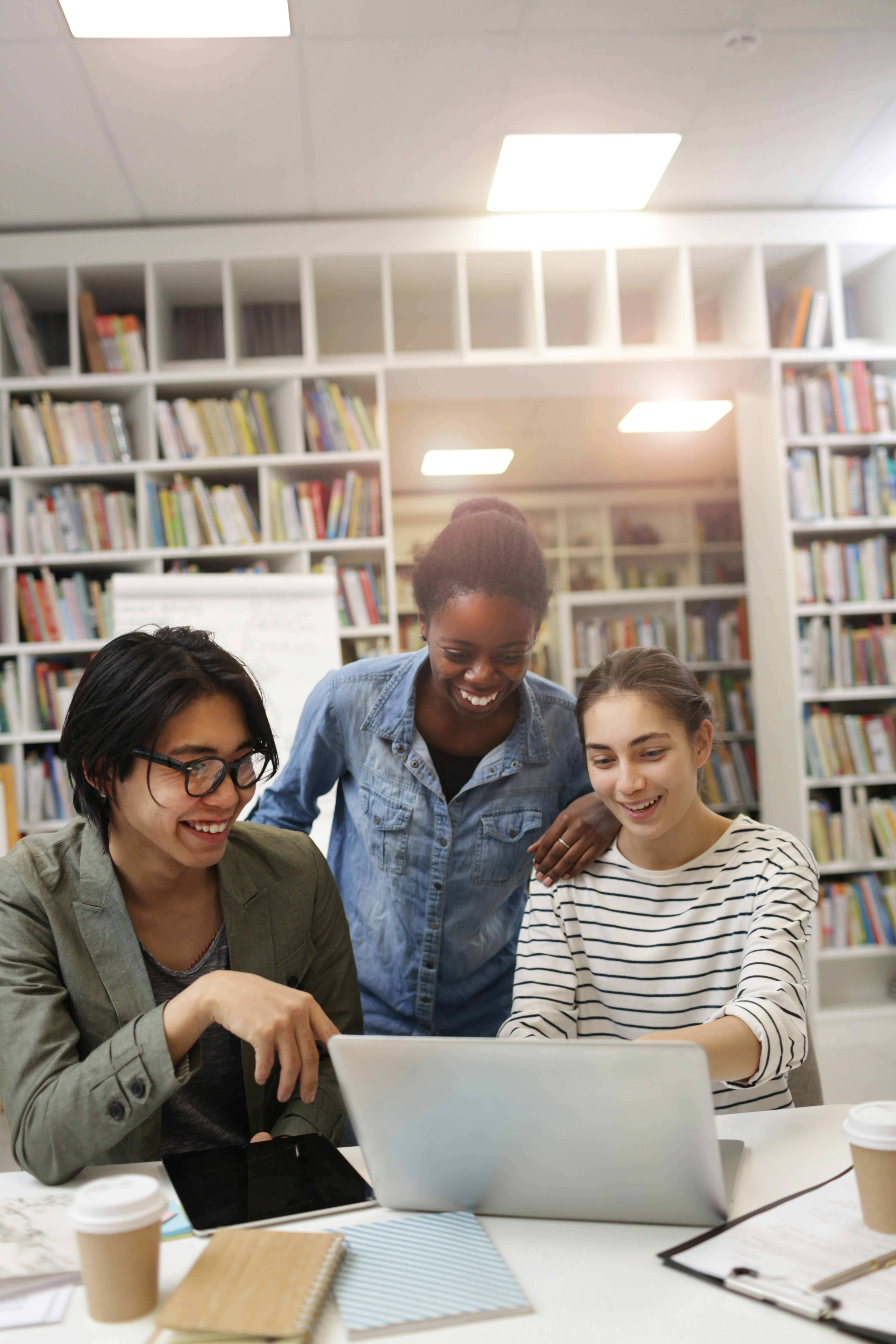 Three colleagues smiling around a laptop in a library workspace